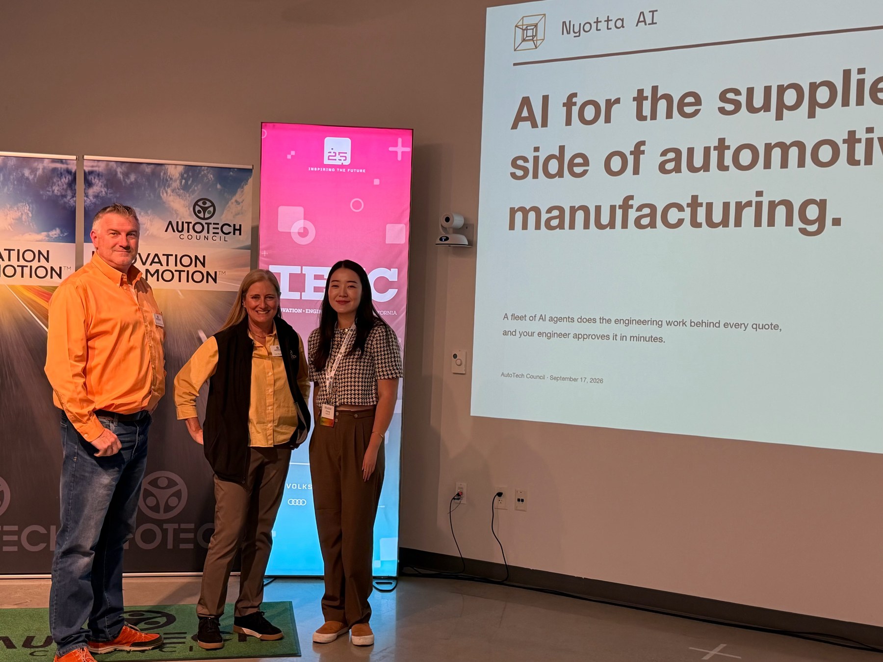 Three attendees at AutoTech Council beside Nyotta AI's presentation on automotive manufacturing, September 17, 2026.